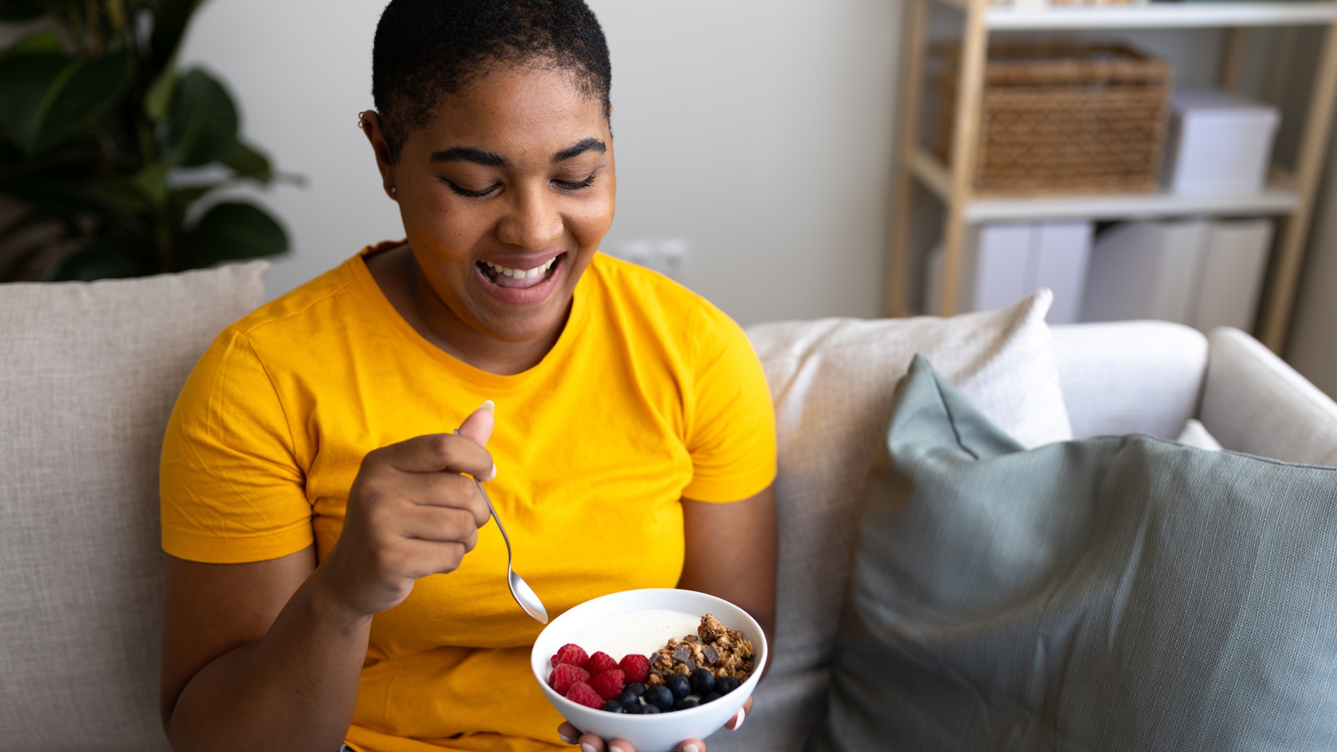 woman in a yellow tshirt sitting on a sofa close to holding a bowl of raspberries,  blueberries and granola. shot from waist up and smiling at her bowl.